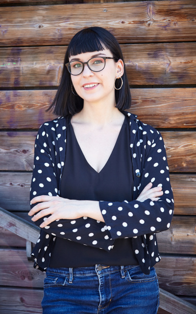 Portrait de Marguerite Pometko, coach en bien-être numérique, les bras croisés, détendue et accoudée sur un mur en bois. Elle porte une veste bleue à pois, un haut noir et un jean.